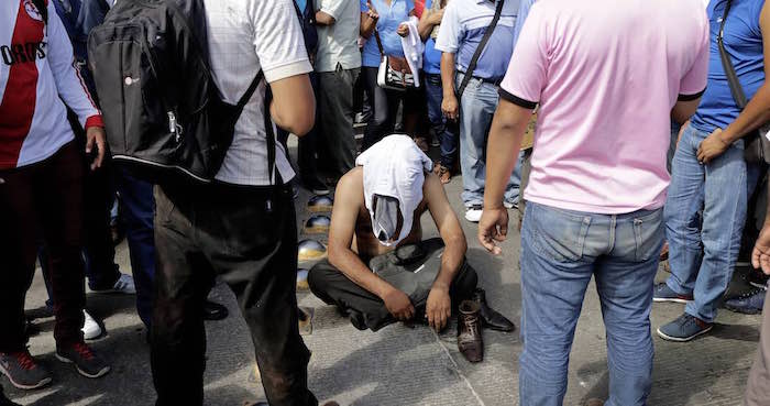 Presunto policía infiltrado sentado en el suelo, con la cara cubierta y sin camiseta, mientras una veintena de manifestantes lo rodean. Foto: Cuartoscuro