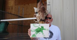 Fotografía del 21 de mayo de 2017, del presidente y propietario del Lion Habitat, Keith Evans, quien posa junto a la jirafa Ozzie y uno de sus dibujos en el interior del santuario animal ubicado a unas 16 millas al sureste de Las Vegas, Nevada (EU). Foto: EFE. 