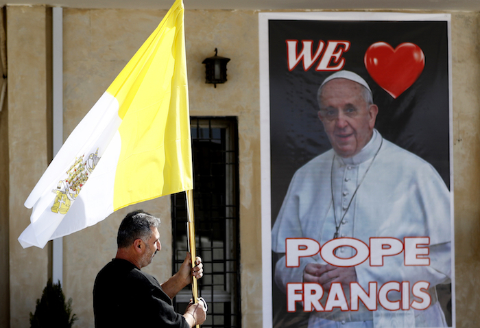 Un sacerdote cristiano sostiene una bandera del vaticano mientras camina ante un cartel del Papa Francisco, durante los preparativos para la visita del papa en Mar Youssif Church en Bagdad, Irak, el viernes 26 de febrero de 2021.