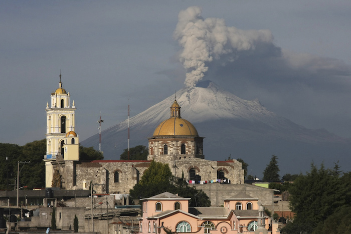 La iglesia principal de San Damián Texoloc, México, al frente del volcán Popocatépetl mientras expulsa cenizas y vapor a inicios de julio de 2013. La sabiduría sagrada de "El Popo" varía de pueblo a pueblo, pero la mayoría coincide en que el volcán no amenaza sus vidas.
